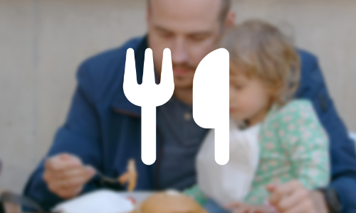 A man feeding a young girl who is sitting on his knee overlayed with an icon of a fork and knife